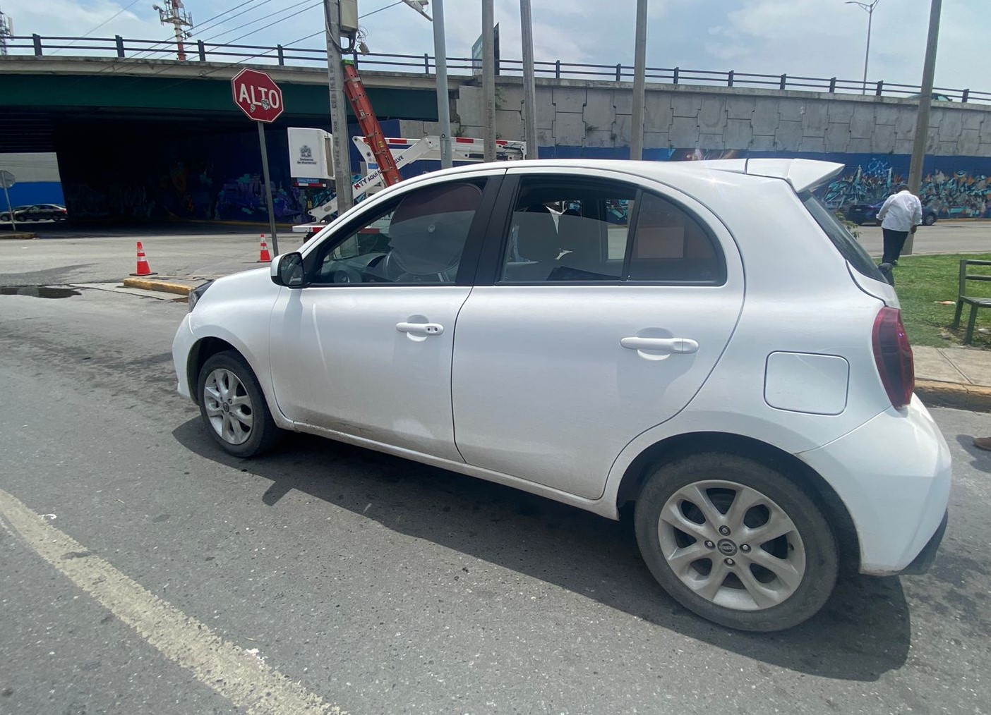 White compact hatchback parked on a city street beside a graffiti wall under an overpass, with traffic cones in the background
