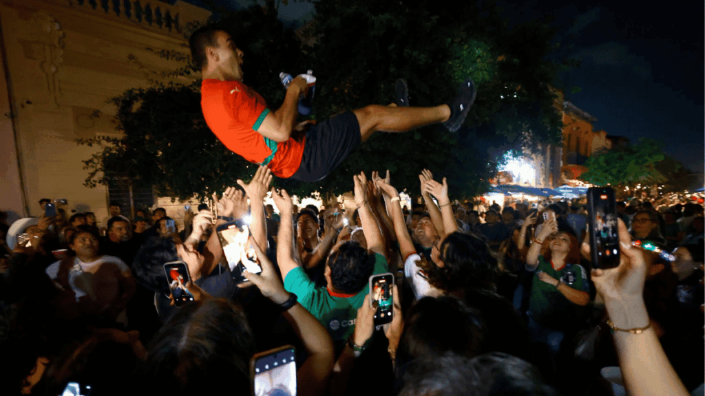 Man in a red shirt crowd-surfing above a cheering crowd at night, with many people holding up smartphones to record the moment.