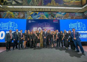Group of about 20 professionals in formal attire posing for a group photo at a 50th anniversary event, with large blue banners that read '50 Aniversario' and 'Gobierno de Monterrey' in the background.