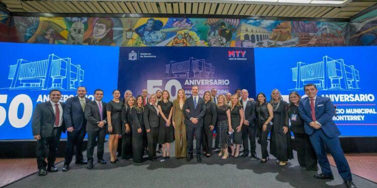 Group of about 20 professionals in formal attire posing for a group photo at a 50th anniversary event, with large blue banners that read '50 Aniversario' and 'Gobierno de Monterrey' in the background.